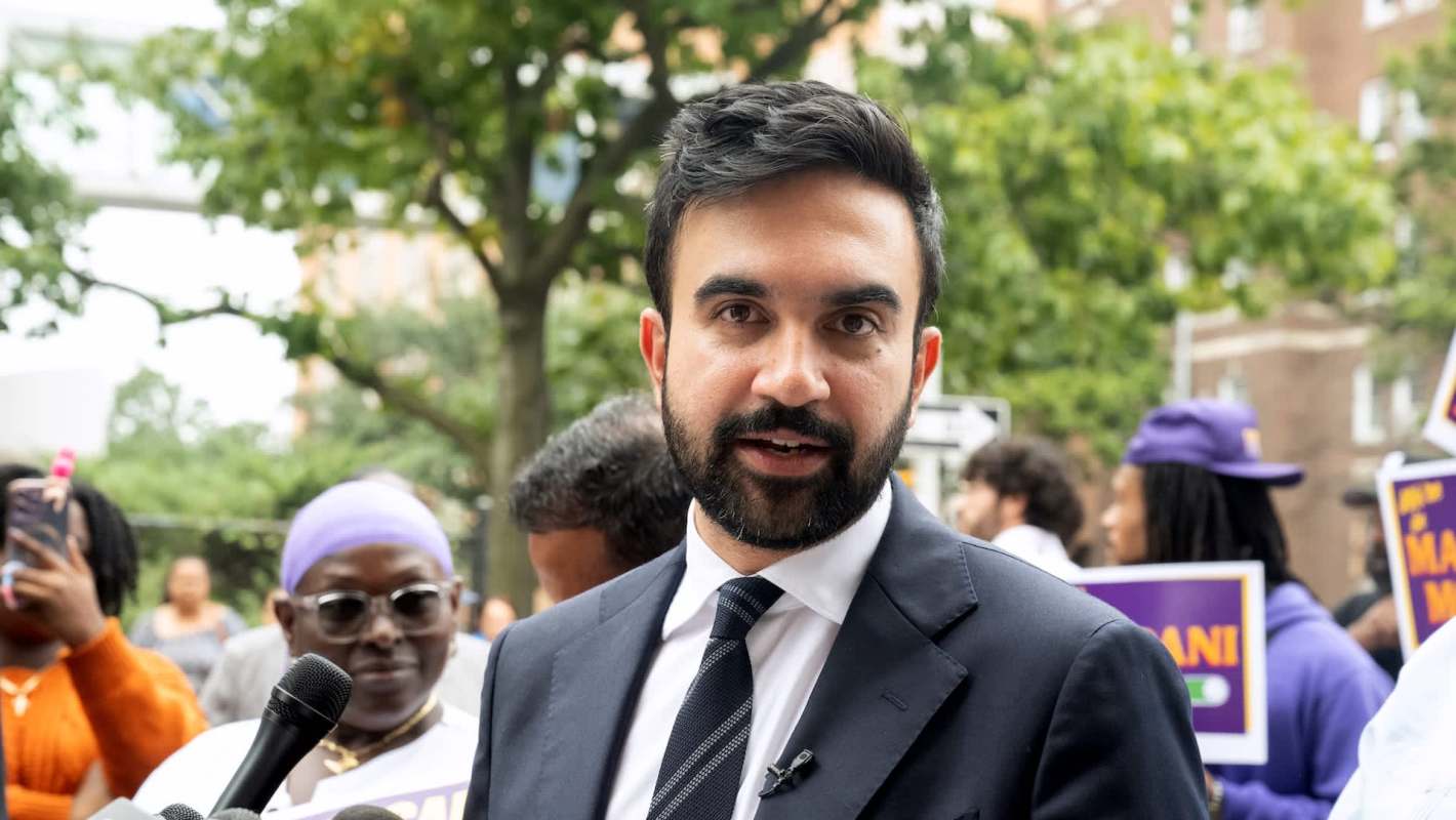 New York City Mayor Zohran Mamdani speaks at an outdoor rally, with trees and supporters in the background.