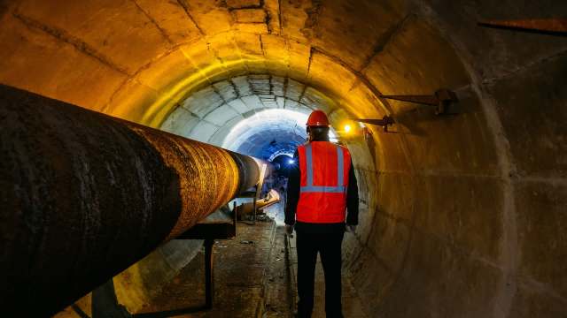 A worker in a safety vest stands in a dimly lit tunnel with large pipes and walls illuminated by warm lights.