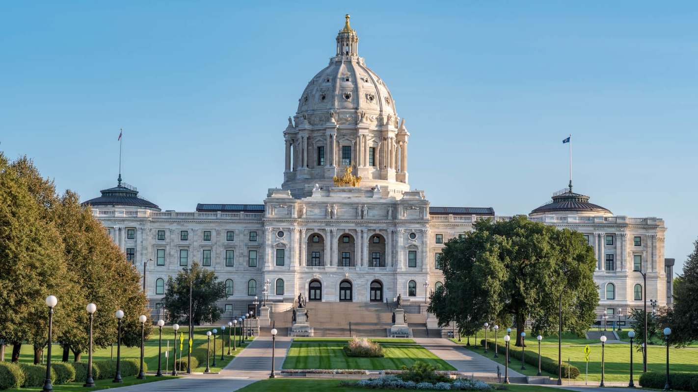 The Minnesota State Capitol building features a prominent dome and elegant stone architecture surrounded by landscaped grounds.