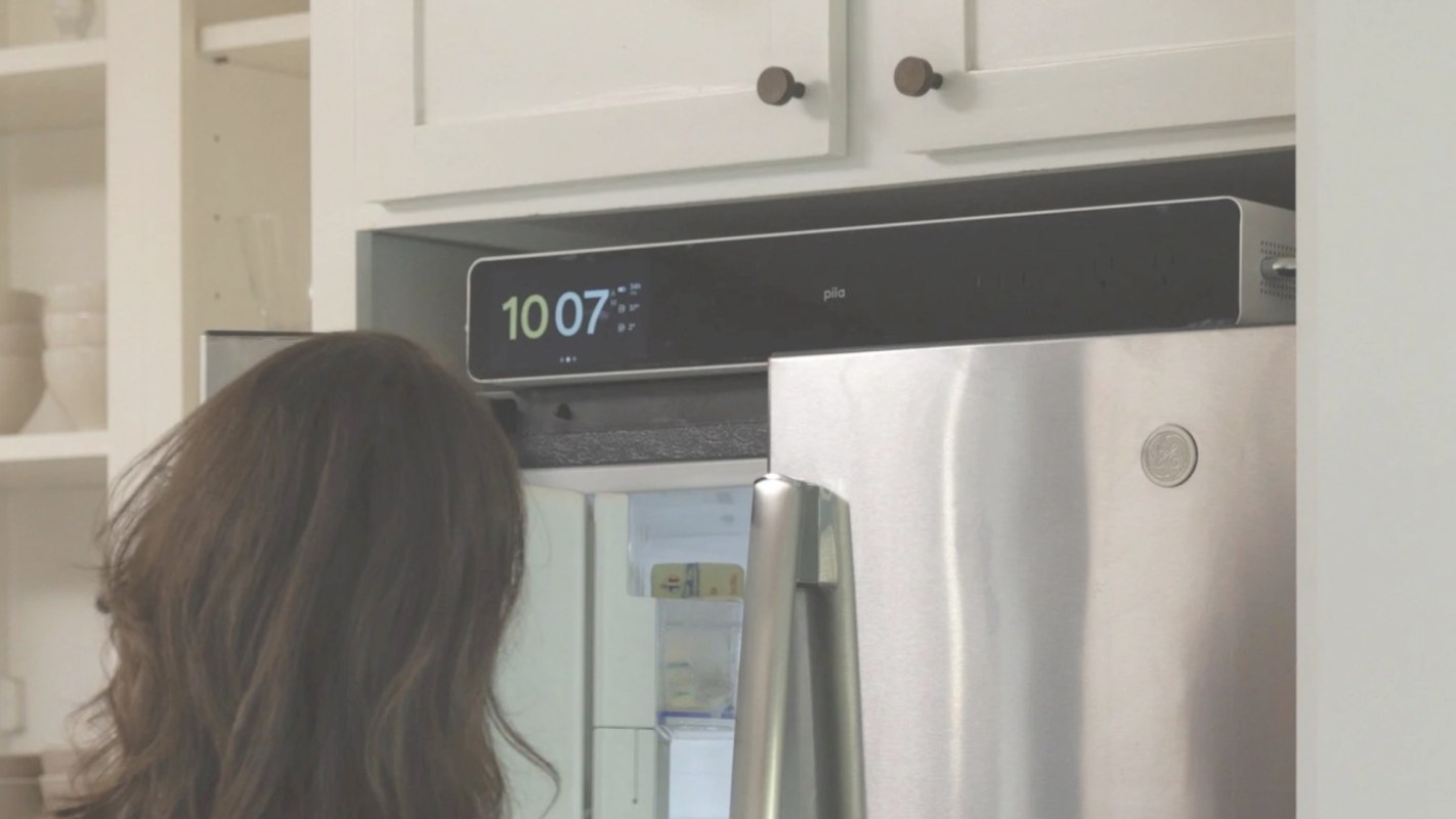 A person stands in front of a refrigerator with a home battery on top of it.