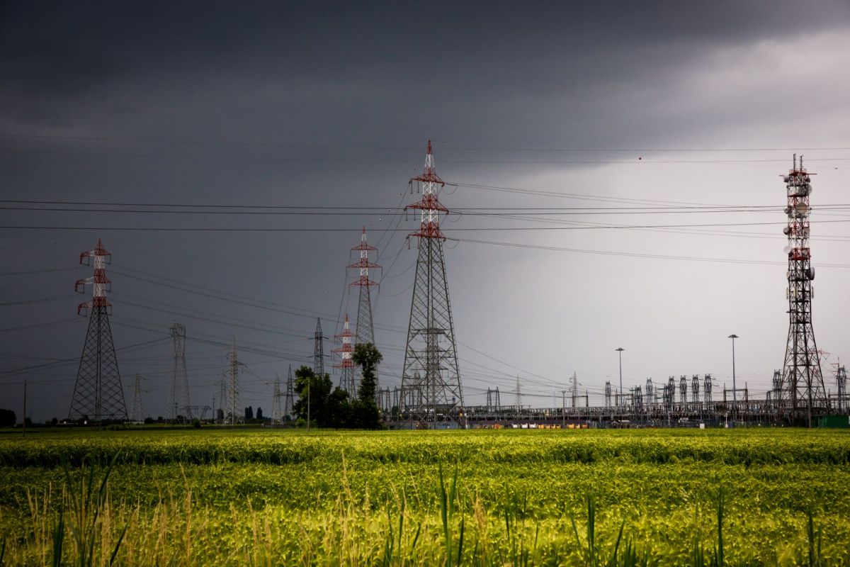 Power lines and tall pylons dominate a rural landscape under a dark, cloudy sky, with green fields in the foreground.