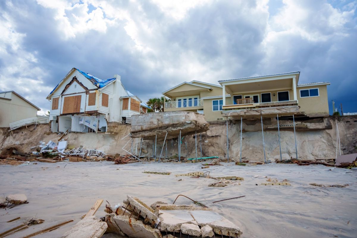 Two houses at the edge of a cliff, one partially collapsed, with debris scattered on the sandy ground below.