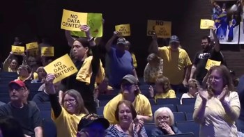 A crowded auditorium with people holding signs that read "NO DATA CENTERS" in protest.