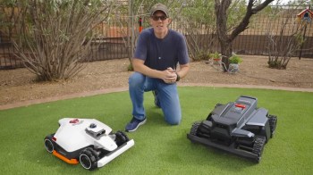 A person kneels on a lawn beside two robotic lawnmowers, with bushes and a fence in the background.