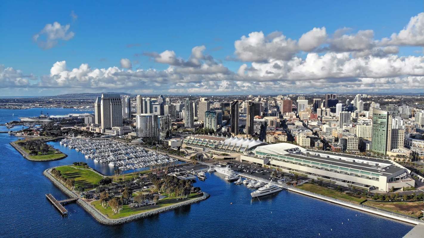 Aerial view of a bustling San Diego skyline with a marina and waterfront park under a vibrant blue sky.