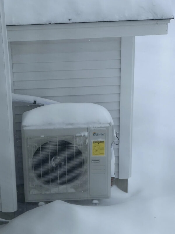 A heat pump covered in snow next to a house's siding on a winter day. 