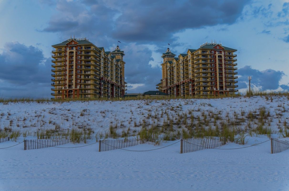 Two tall beachside buildings rise above a dune of white sand under a cloudy blue sky.
