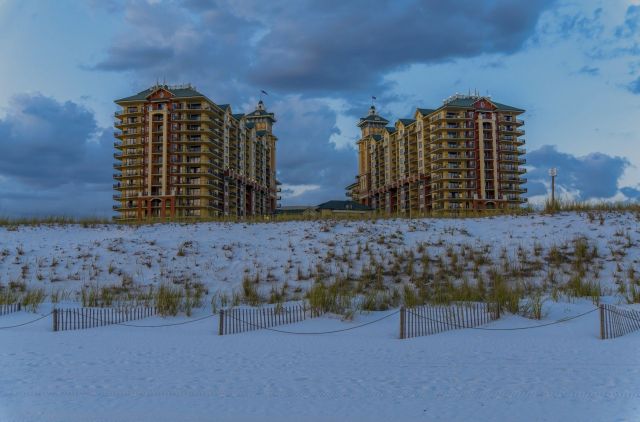 Two tall beachside buildings rise above a dune of white sand under a cloudy blue sky.