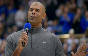 Shane Battier speaks into a microphone during a Duke basketball event, wearing a gray shirt and engaging the audience.