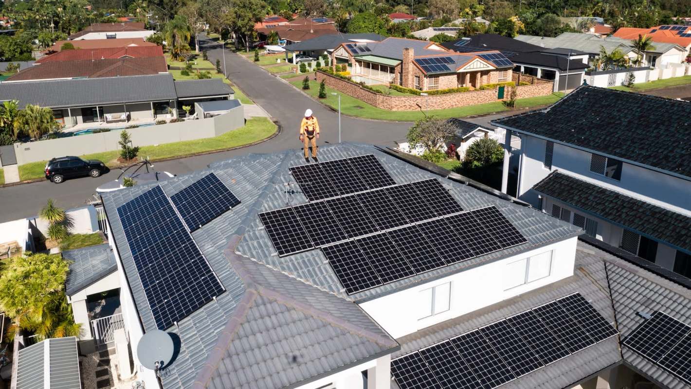A worker stands on a roof covered with solar panels in a residential neighborhood.