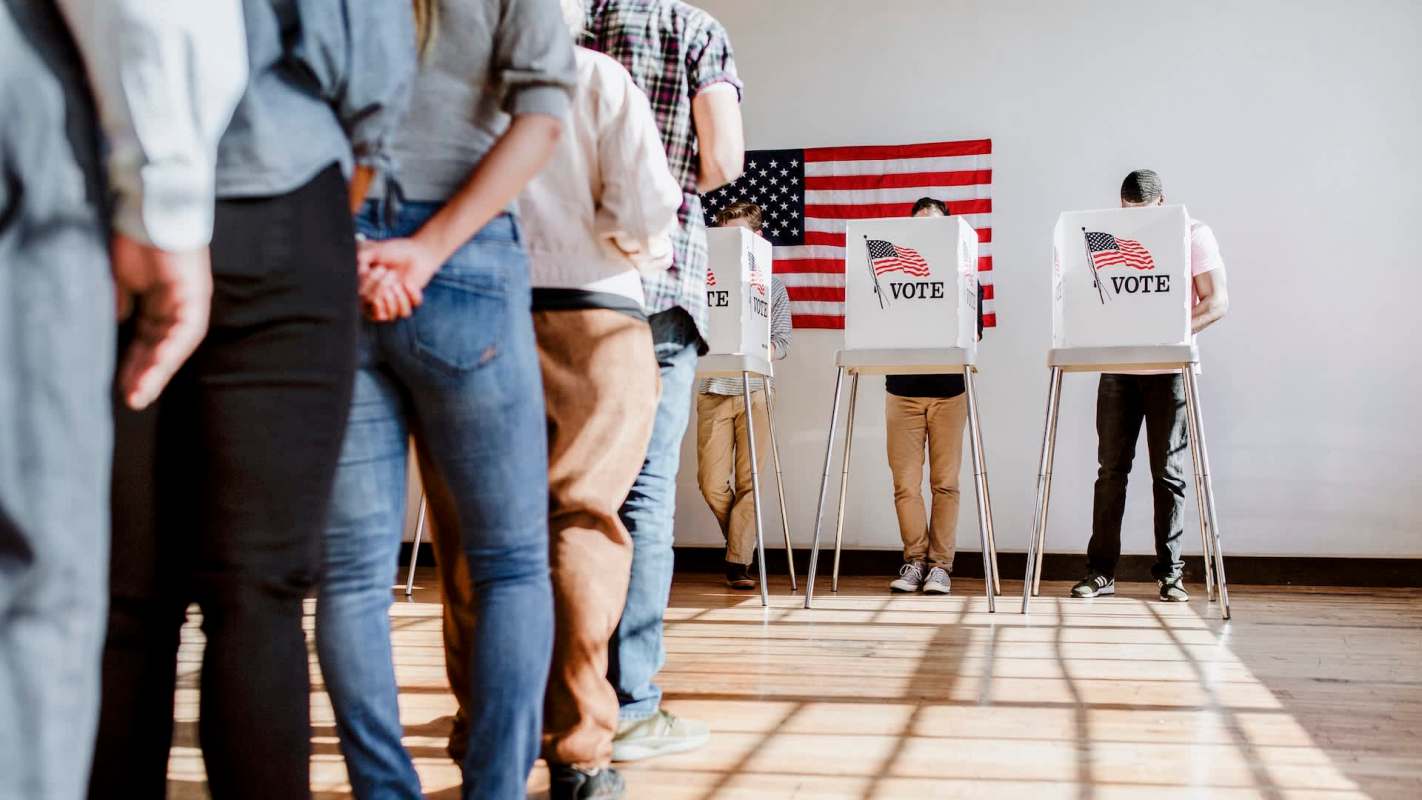 A line of voters casting ballots at voting booths with an American flag in the background.