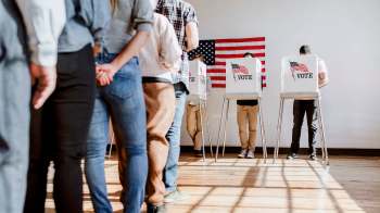 A line of voters casting ballots at voting booths with an American flag in the background.