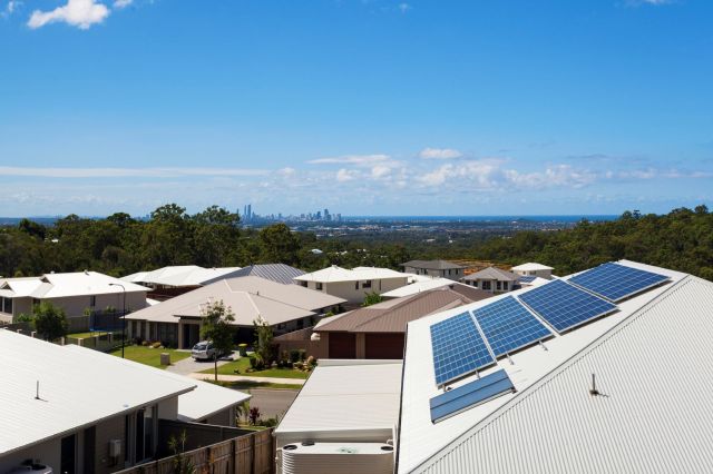 A view of suburban rooftops with solar panels and a skyline in the distance under a clear blue sky.