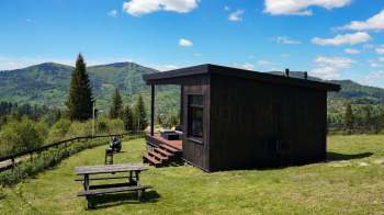 A modern black cabin with a porch, situated on a green hillside with mountains in the background.