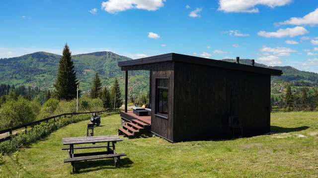 A modern black cabin with a porch, situated on a green hillside with mountains in the background.