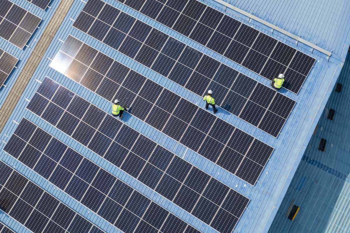 Workers in safety gear are installing solar panels on a blue rooftop.