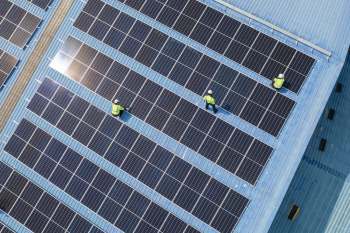Workers in safety gear are installing solar panels on a blue rooftop.