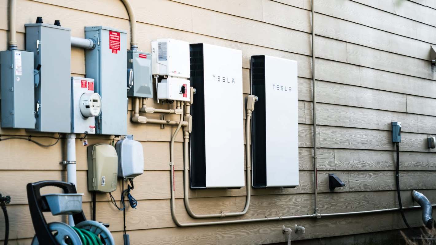 Two Tesla battery units mounted on a beige wall, alongside various electrical meters and connections.