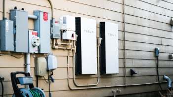 Two Tesla battery units mounted on a beige wall, alongside various electrical meters and connections.