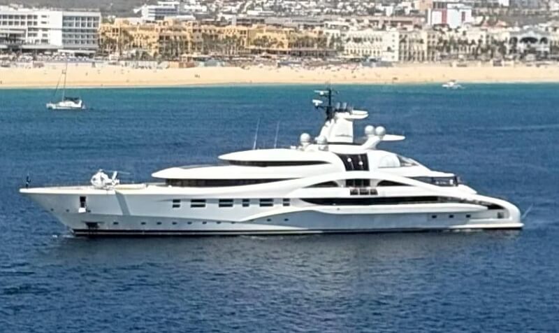 A sleek white yacht floating on blue waters in front of a beach and cityscape.  