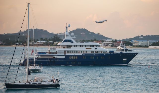 A luxury yacht sails in a harbor while a plane ascends in the background against a pastel sky.