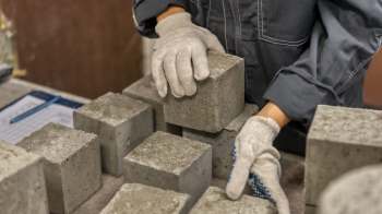 A person in gloves is stacking concrete blocks on a workbench with a clipboard nearby.