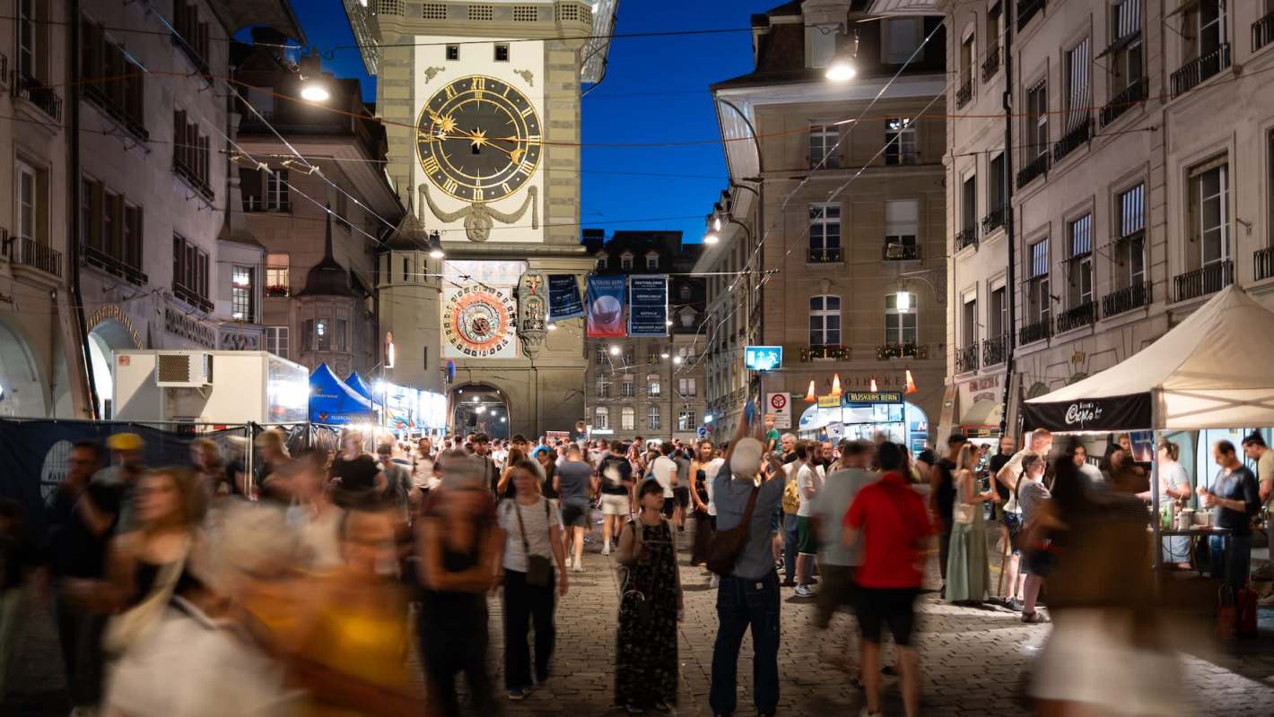 A bustling street market at night with a large clock tower in the background and crowds of people.