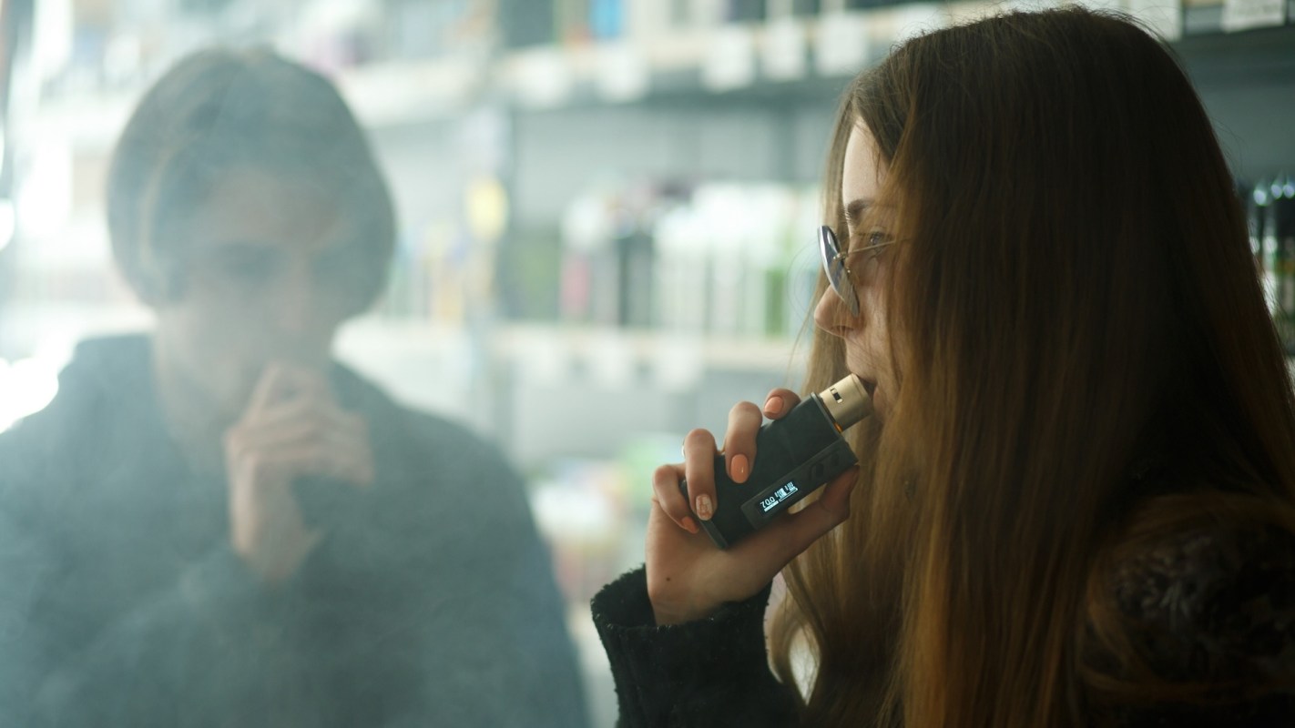 A young woman vapes while a blurred figure of a man watches in a smoke-filled environment.