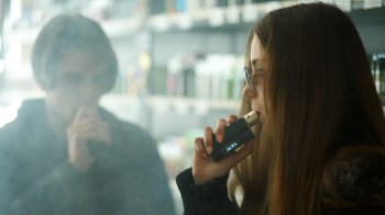 A young woman vapes while a blurred figure of a man watches in a smoke-filled environment.