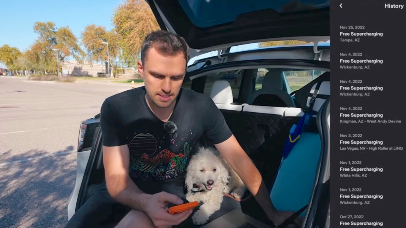 A man sits in the back of his Tesla with a small dog, looking at his phone while parked in a sunny, open area.