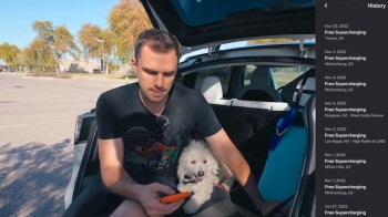 A man sits in the back of his Tesla with a small dog, looking at his phone while parked in a sunny, open area.