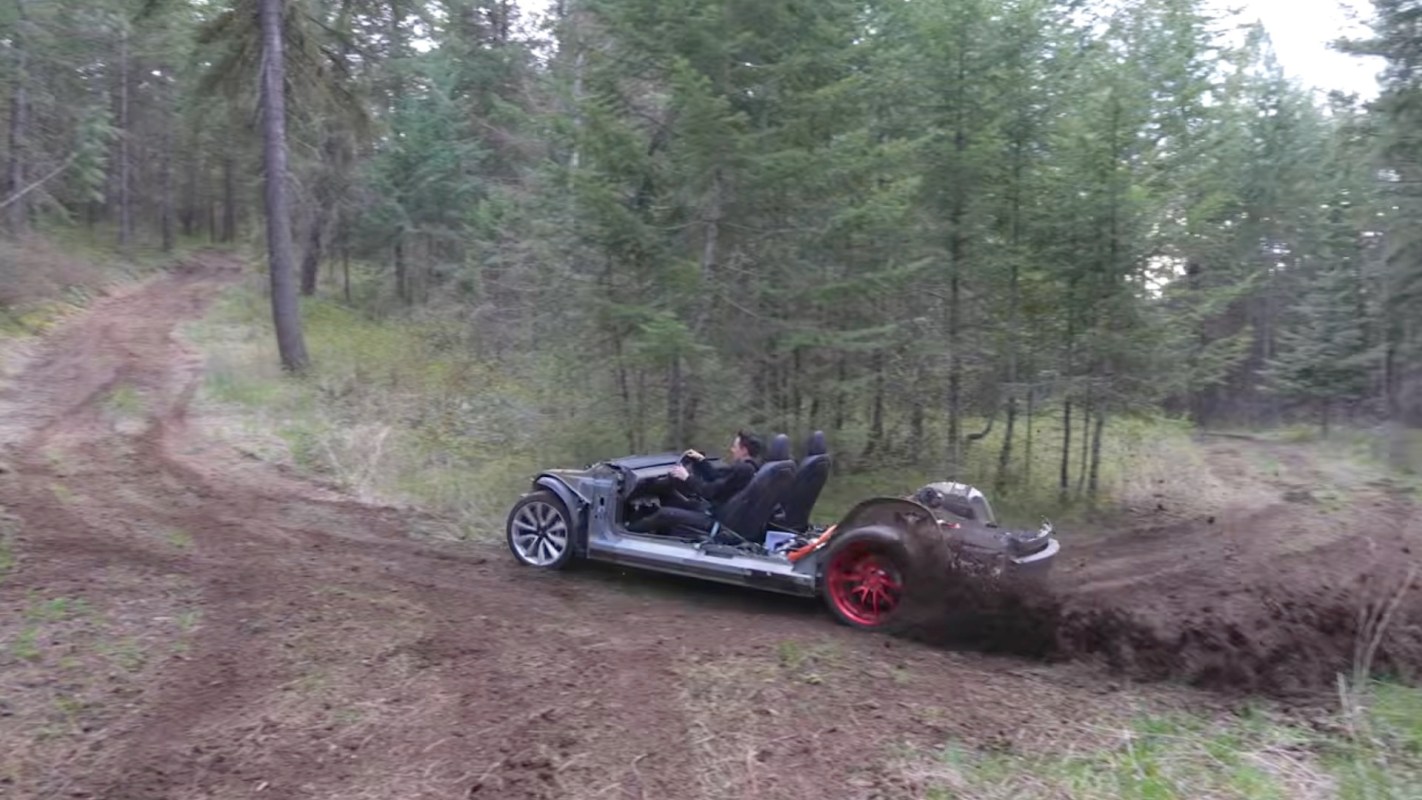 A partially assembled Tesla car drifts on a muddy forest trail, kicking up dirt and debris.