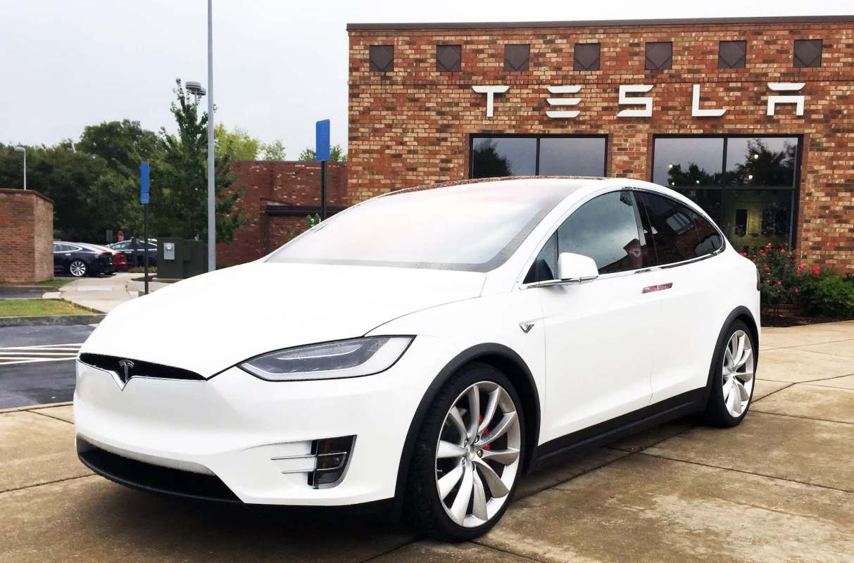 A white Tesla Model X EV in front of a building with the Tesla logo on it.