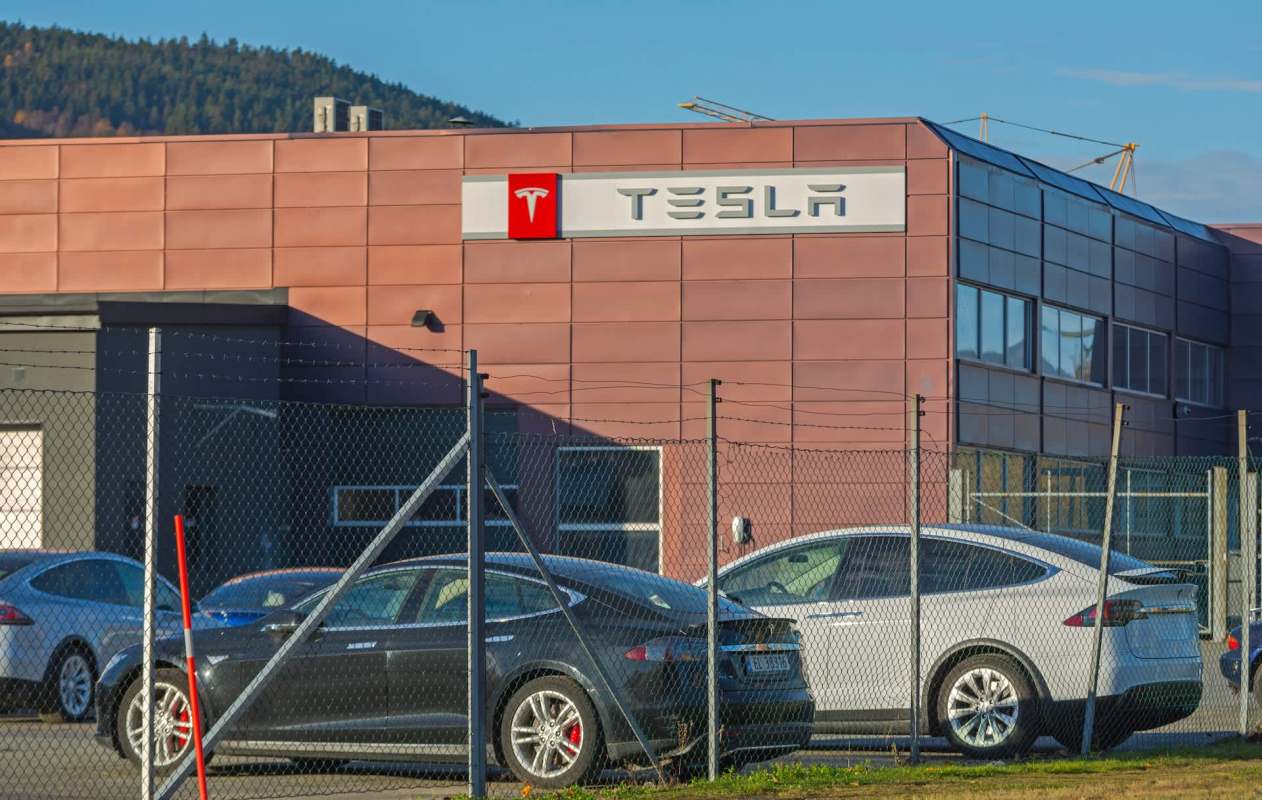A Tesla dealership with several parked cars behind a fence and a mountain in the background.