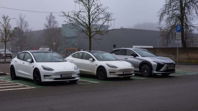 Three Tesla electric cars parked in a lot on a foggy day, with trees and buildings in the background.