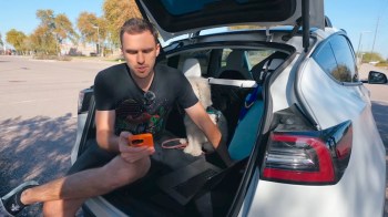 A man sits in the back of his white Tesla Model Y, using a phone, with a small dog beside him.