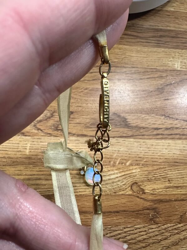 A hand holds a delicate ribbon necklace with a gold pendant featuring blue and white gemstones.