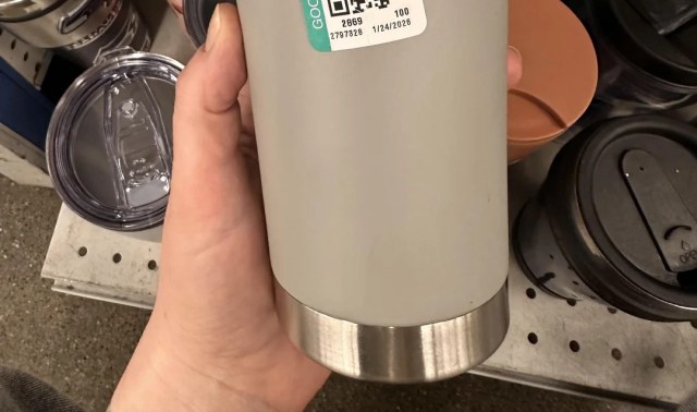 A person holding a gray tumbler with a stainless steel bottom and a price tag, surrounded by various containers.