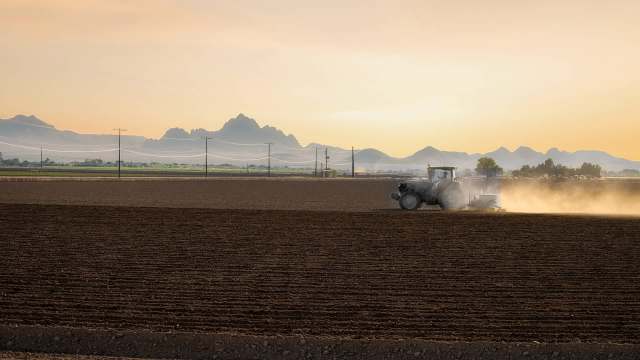 A tractor plows a dusty field against a backdrop of mountains and a sunset sky.