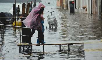 A person in a pink poncho helps a child navigate floodwaters on a raised platform beside a marina in Venice, Italy.