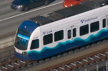 A Sound Transit Link light rail train travels along the tracks on a floating bridge beside a Seattle highway.