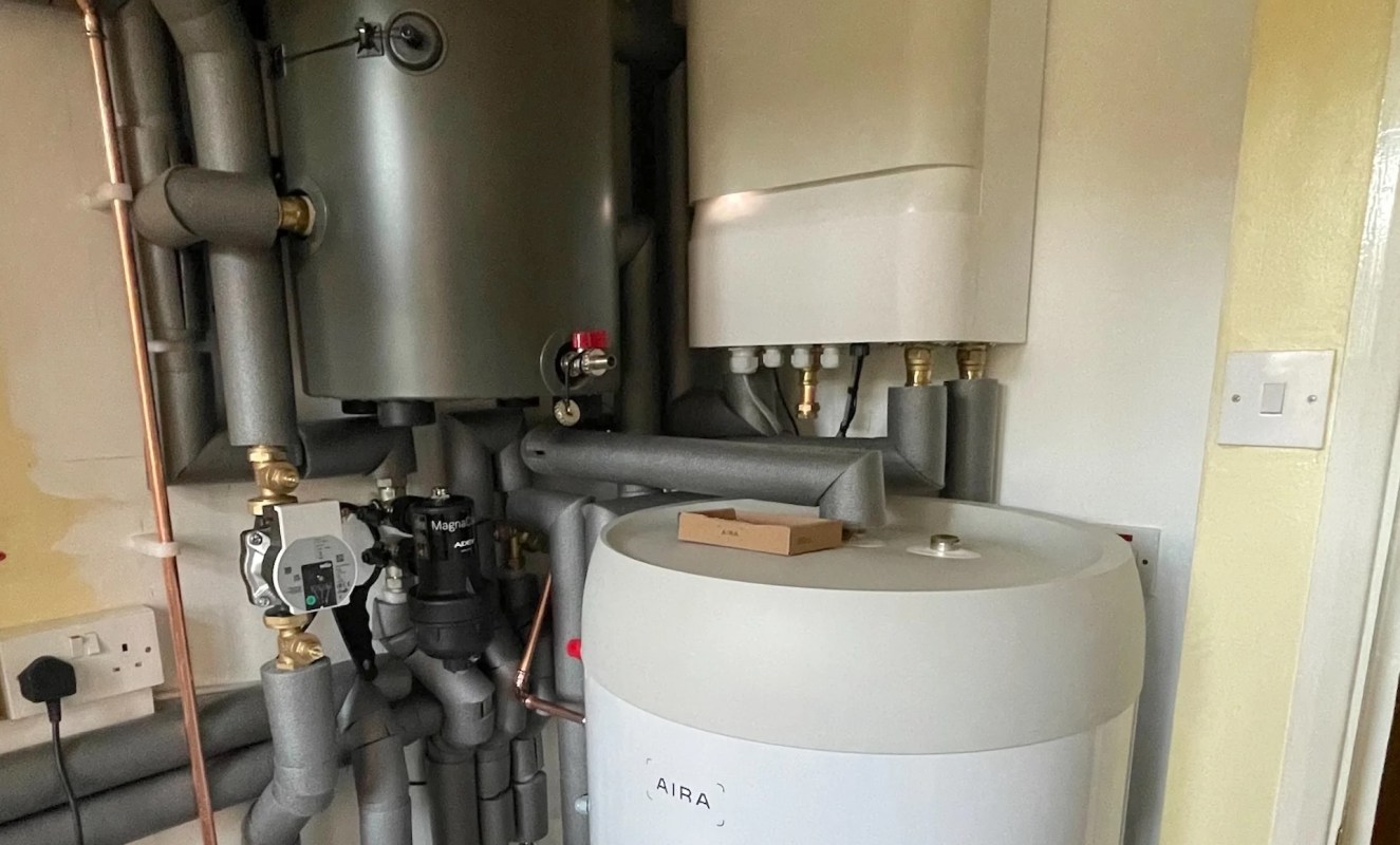 A series of pipes and valves connected to a gray water heater and a white storage tank on a wall.