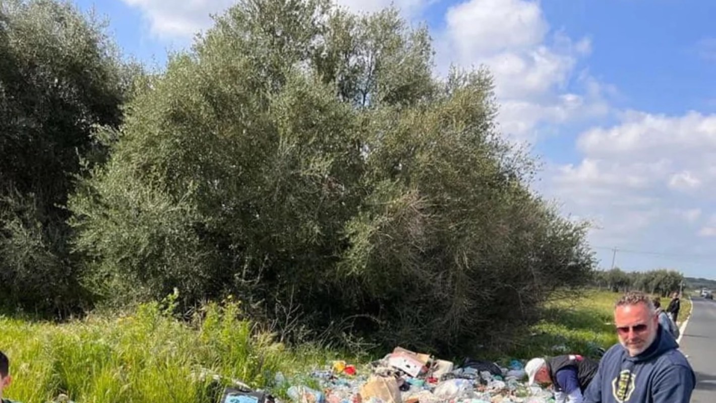 A group of people cleaning up garbage along a roadside, surrounded by greenery and a clear blue sky.