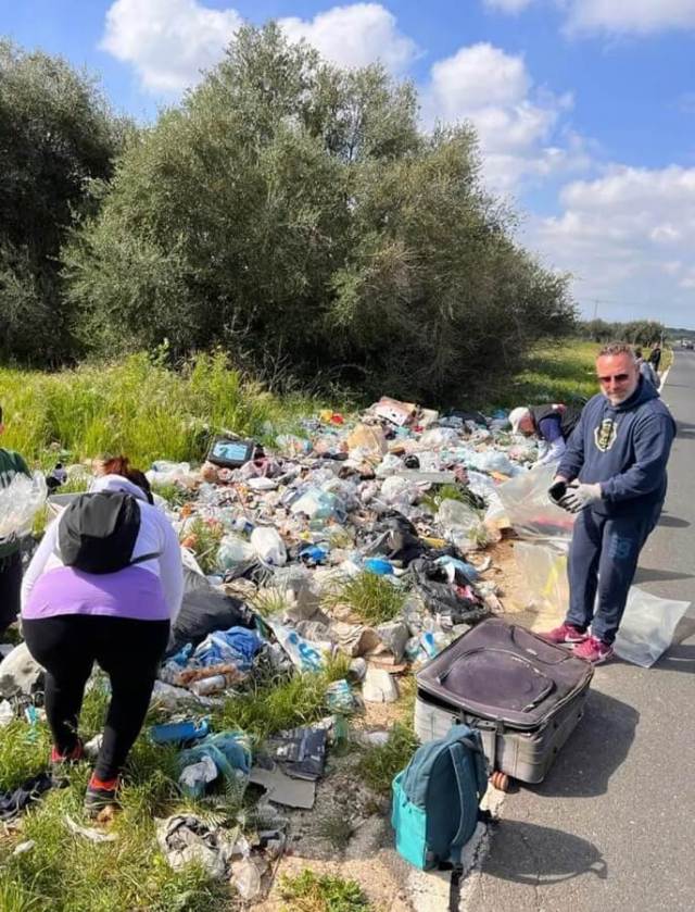 A group of people cleaning up garbage along a roadside, surrounded by greenery and a clear blue sky.