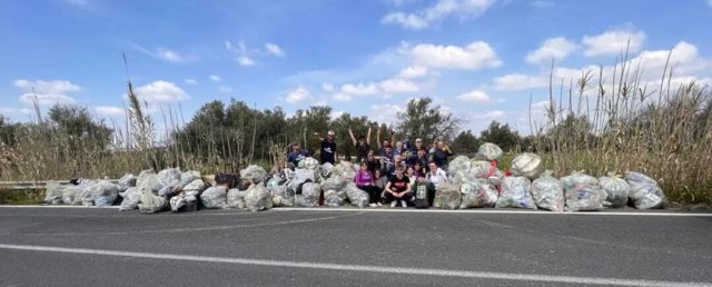 A group of people surrounding multiple bags or garbage along a roadside, surrounded by greenery and a clear blue sky.