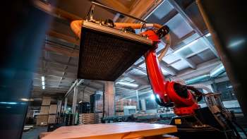 A red robotic arm lifts a flat surface in a warehouse filled with stacked pallets and machinery.
