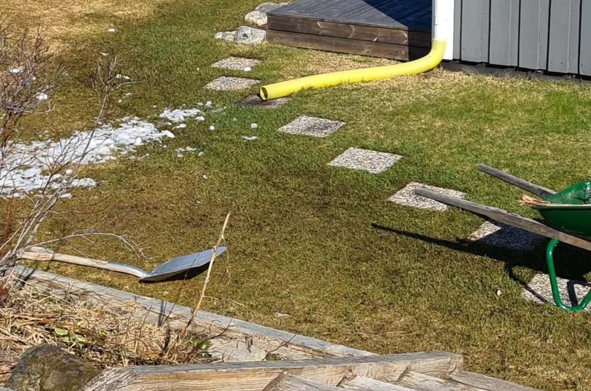A garden scene featuring a shovel, a wheelbarrow, stepping stones, and patches of snow on green grass.