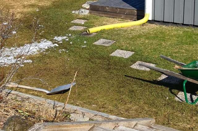 A garden scene featuring a shovel, a wheelbarrow, stepping stones, and patches of snow on green grass.