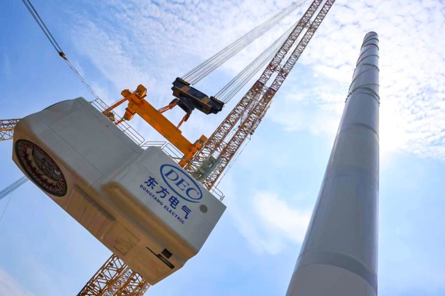 A crane lifting a large turbine component against a bright blue sky with faint clouds.