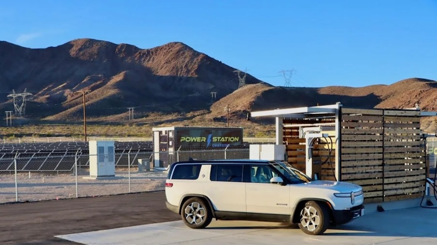 A white electric vehicle is parked near a charging station with solar panels and mountains in the background.
